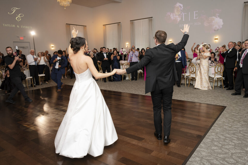 Bride and groom enter a wedding reception, holding hands and waving to cheering guests in a decorated hall.