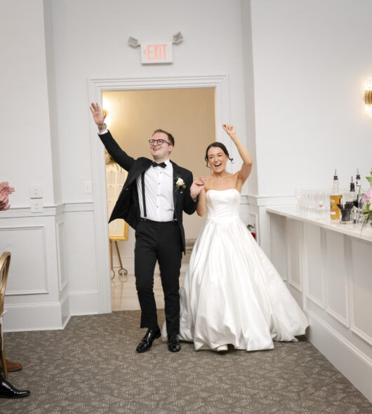 A newlywed couple enters a reception, smiling and waving as guests applaud and cheer.