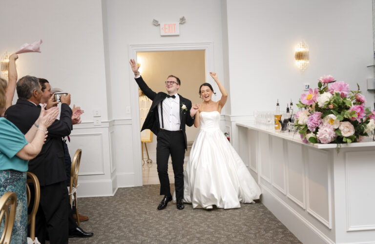 A newlywed couple enters a reception, smiling and waving as guests applaud and cheer.