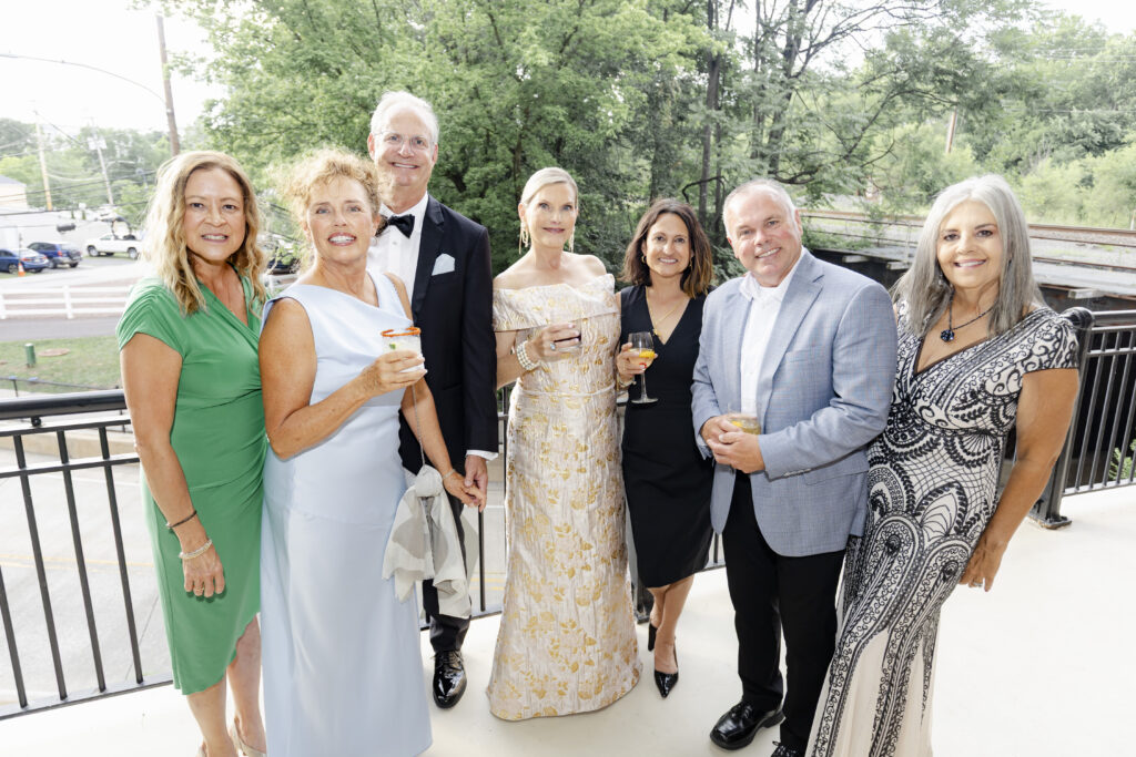 Seven adults dressed formally, smiling and holding drinks, pose together on an outdoor balcony with trees in the background.