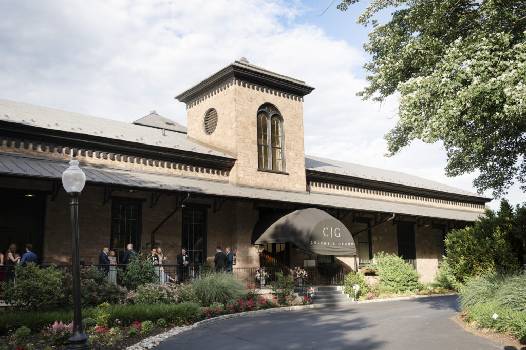 Columbia Station event venue with people mingling outside, awning entrance, greenery, and blue sky.