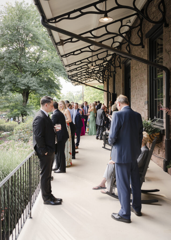 People in formal attire gather and converse on a covered outdoor veranda with trees and greenery in the background.