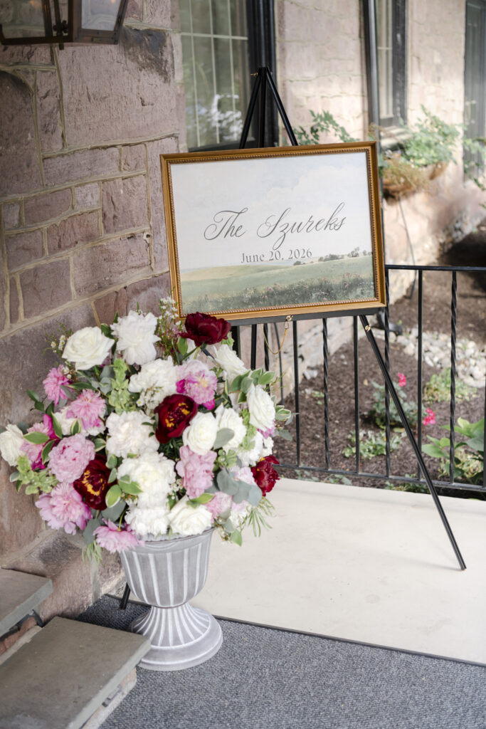 A floral arrangement sits below a framed sign reading “The Szureks, June 20, 2026” on an easel by a stone wall.