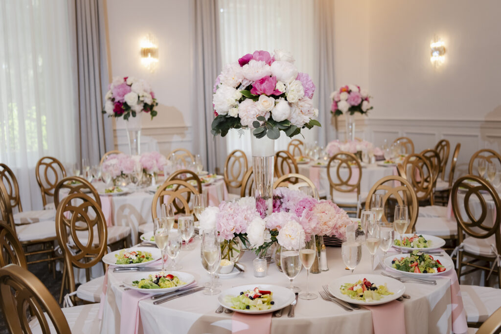 Elegant wedding reception with round tables, pink and white flowers, gold chairs, and salads set for guests.