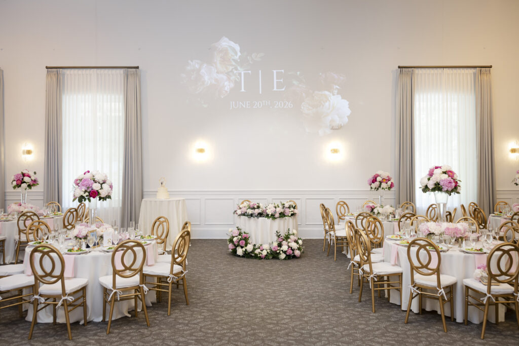 Elegant wedding reception hall with round tables, pink floral centerpieces, and gold chairs, wedding date on wall.